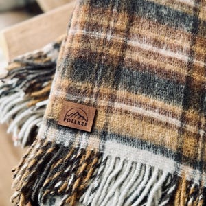 May include: A cozy plaid blanket in shades of brown, tan, and gray with a fringed edge. The blanket features a leather patch with the text "FOLLKEE" and a mountain graphic. The blanket is folded, showcasing its texture and pattern.