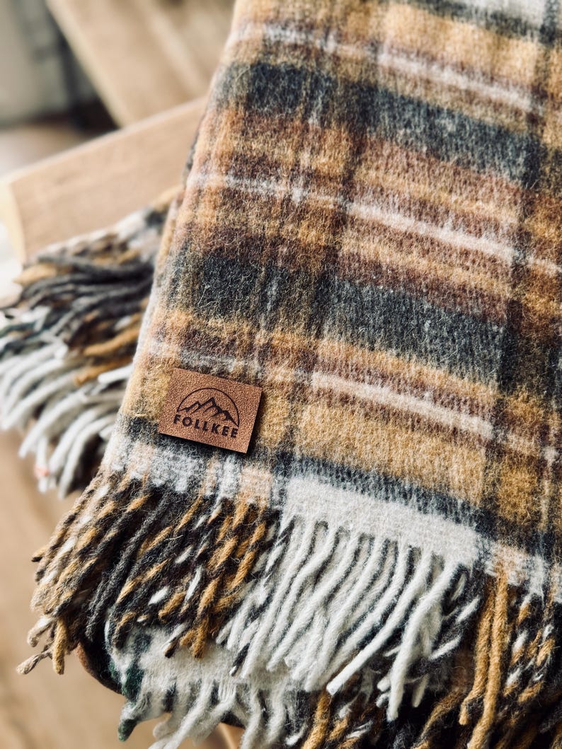 May include: A cozy plaid blanket in shades of brown, tan, and gray with a fringed edge. The blanket features a leather patch with the text "FOLLKEE" and a mountain graphic. The blanket is folded, showcasing its texture and pattern.
