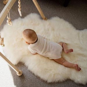 May include: A baby in a white onesie lies on a fluffy, off-white sheepskin rug. A wooden baby gym with hanging wooden toys is partially visible in the upper left corner. The rug is on a gray carpet.