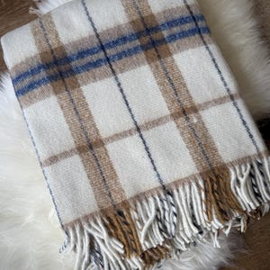 May include: A cream-colored plaid blanket with brown and blue stripes. The blanket has a fringed edge in shades of brown and cream. The blanket is folded and resting on a white, fluffy surface.