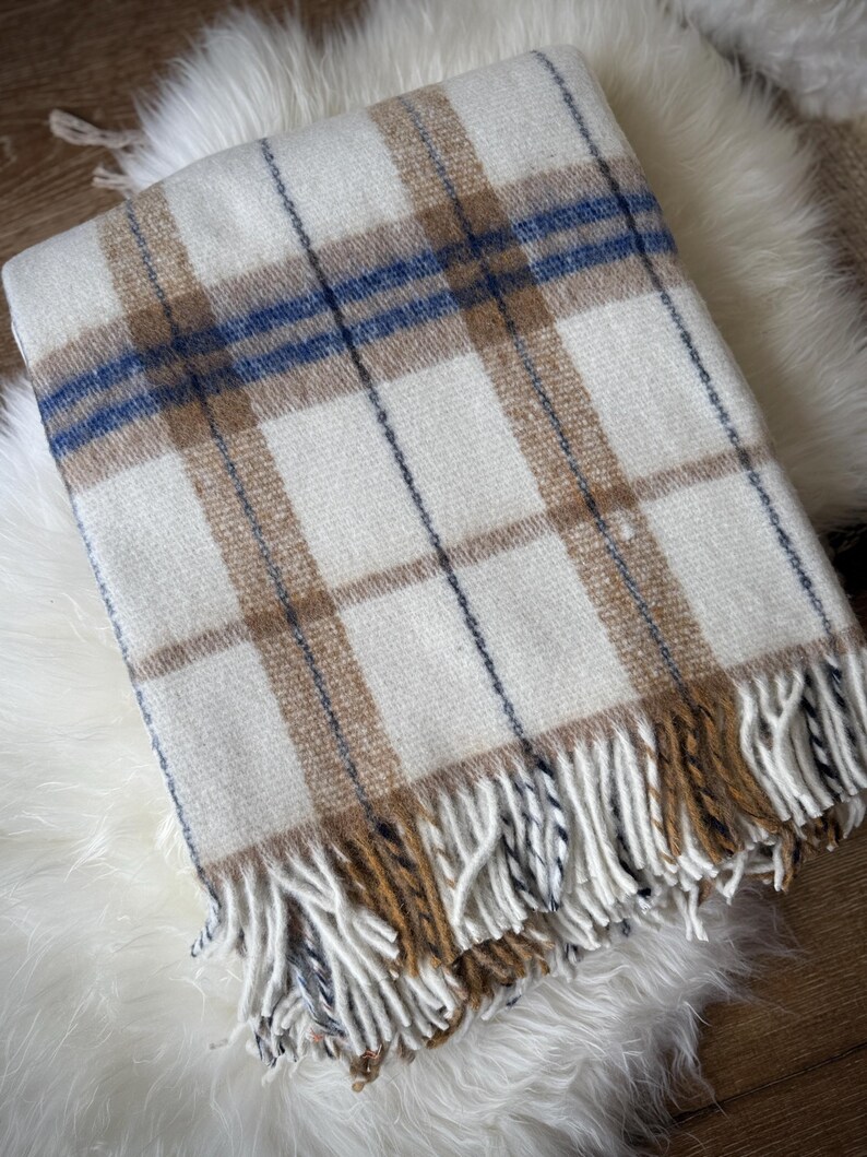 May include: A cream-colored plaid blanket with brown and blue stripes. The blanket has a fringed edge in shades of brown and cream. The blanket is folded and resting on a white, fluffy surface.