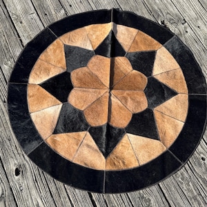 May include: Round cowhide rug with a geometric design. The rug features a central starburst pattern in shades of tan and black, surrounded by a black border. The rug is made of natural materials and has a rustic aesthetic.