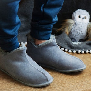 May include: Grey suede slippers with a fleece lining. The slippers have a rounded toe and a low heel.