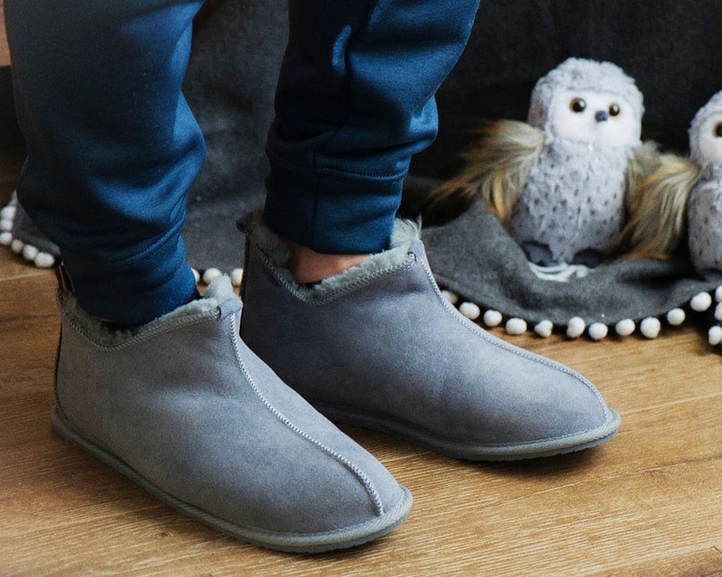 May include: Grey suede slippers with a fleece lining. The slippers have a rounded toe and a low heel.