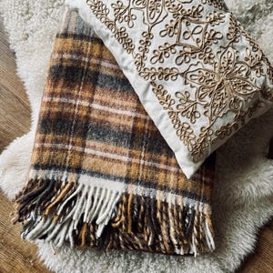 May include: A cozy arrangement featuring a plaid blanket with fringe in shades of brown, tan, and white, paired with a white pillow adorned with gold embroidery. The items rest on a fluffy, white rug.