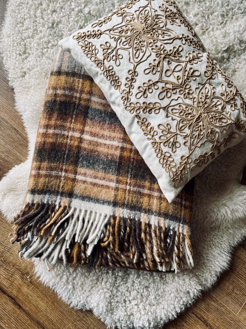 May include: A cozy arrangement featuring a plaid blanket with fringe in shades of brown, tan, and white, paired with a white pillow adorned with gold embroidery. The items rest on a fluffy, white rug.