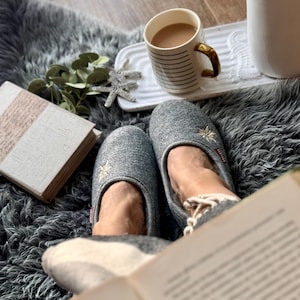 Pode incluir: Uma cena aconchegante com uma x&iacute;cara de caf&eacute;, um livro e um par de pantufas cinzas com flores bordadas. As pantuflas est&atilde;o em um tapete cinza felpudo.