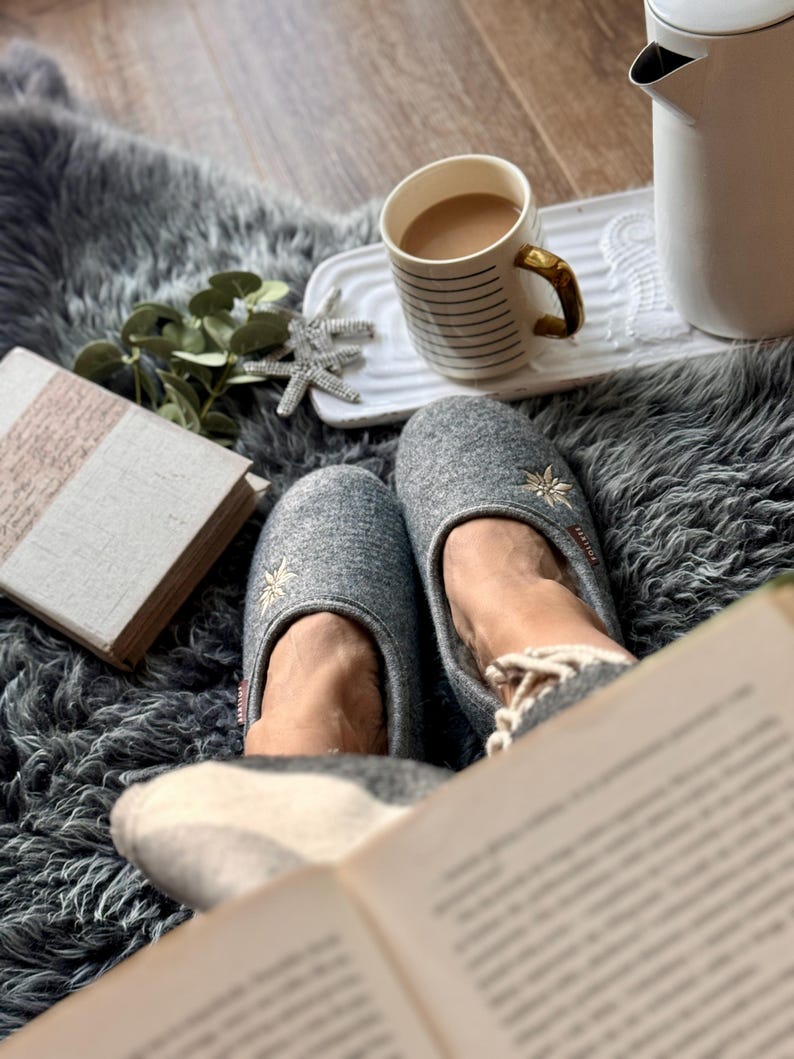 Pode incluir: Uma cena aconchegante com uma x&iacute;cara de caf&eacute;, um livro e um par de pantufas cinzas com flores bordadas. As pantuflas est&atilde;o em um tapete cinza felpudo.