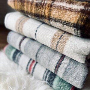 May include: A stack of folded blankets with various plaid patterns. The top blanket is brown and tan, followed by white, gray, and green blankets. The blankets are soft and cozy, perfect for adding warmth and style to any room.