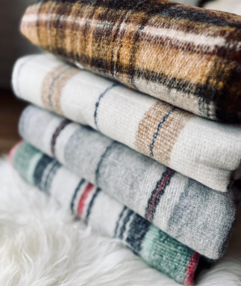 May include: A stack of folded blankets with various plaid patterns. The top blanket is brown and tan, followed by white, gray, and green blankets. The blankets are soft and cozy, perfect for adding warmth and style to any room.