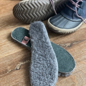May include: A pair of gray fleece shoe insoles with a black felt backing. The insoles are folded in half and the fleece side is facing the camera.