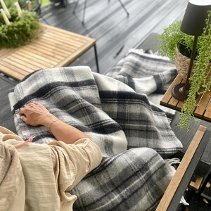 May include: A gray and white plaid blanket draped over a dark surface. The blanket has a fringe along the edge. A person is sitting on the surface, wearing a beige top. A small table with a lamp and a plant is in the background.