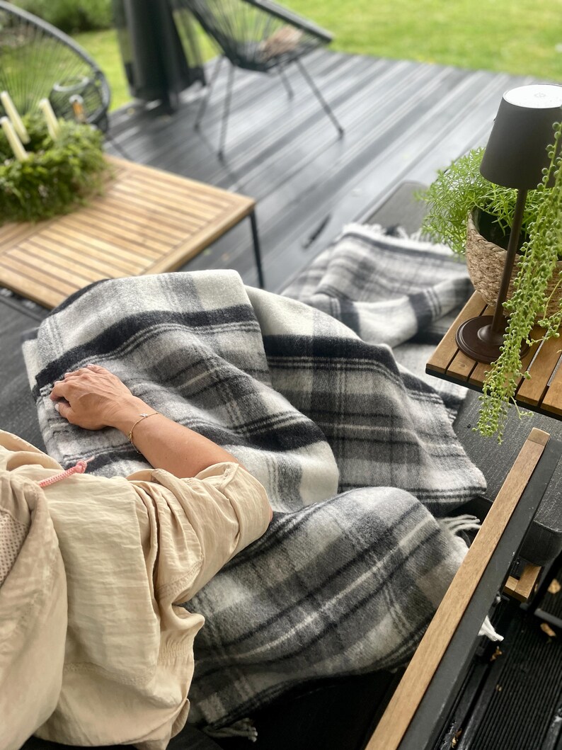 May include: A gray and white plaid blanket draped over a dark surface. The blanket has a fringe along the edge. A person is sitting on the surface, wearing a beige top. A small table with a lamp and a plant is in the background.