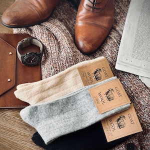 May include: Three pairs of socks in beige, gray, and black, folded on a brown wooden surface. Each pair has a brown paper tag with the word "GOAT" and "WOOL" printed on it. The tags also list the sizes for women and men.