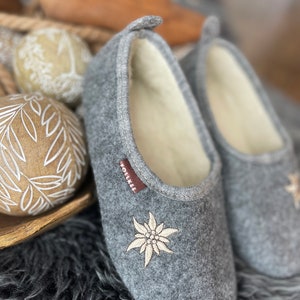 Pode incluir: Um par de pantufas de l&atilde; cinzas com uma flor de edelweiss branca bordada. As pantufas t&ecirc;m um forro de l&atilde; branca e uma etiqueta marrom "Follkee" no lado.