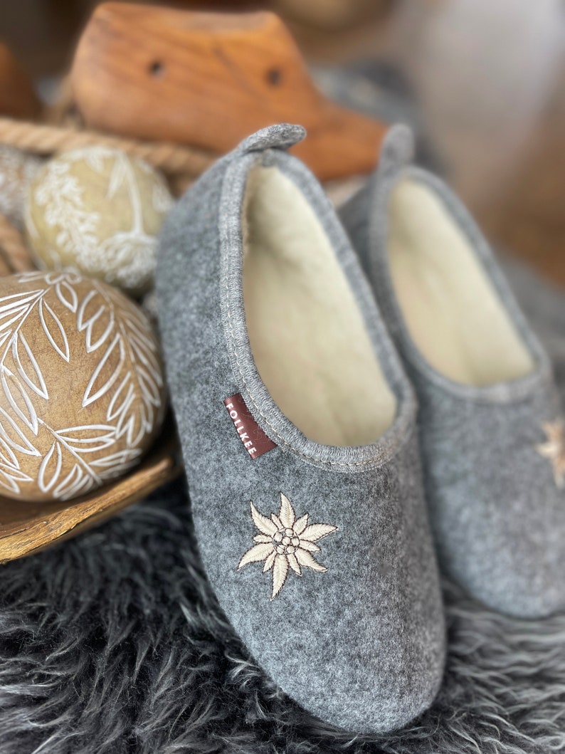 Pode incluir: Um par de pantufas de l&atilde; cinzas com uma flor de edelweiss branca bordada. As pantufas t&ecirc;m um forro de l&atilde; branca e uma etiqueta marrom "Follkee" no lado.