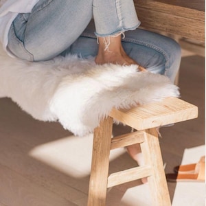 May include: A small, rectangular wooden bench with four angled legs and a natural light wood finish. The top of the bench is covered with a thick, plush white faux fur throw, providing a soft texture that contrasts with the solid wood structure.