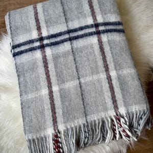 May include: A folded gray plaid blanket with white, navy, and red accents. The blanket has a fringed edge and is displayed on a white, fluffy surface. The plaid pattern is a classic design, suitable for home decor.