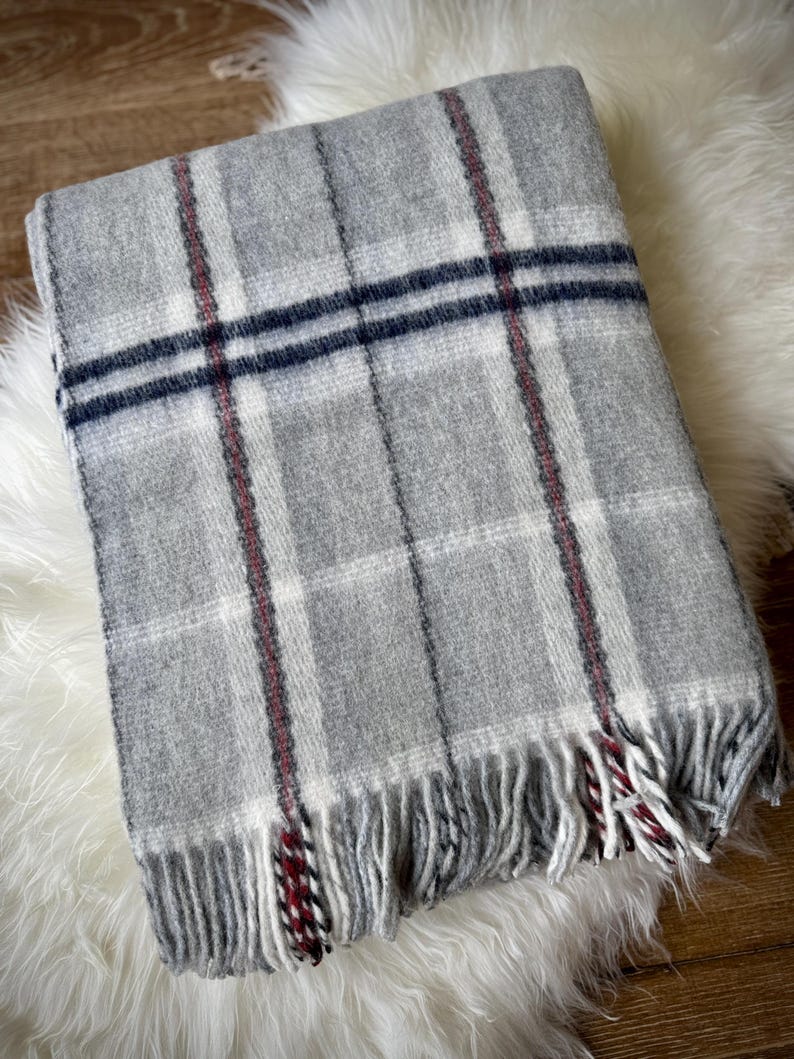 May include: A folded gray plaid blanket with white, navy, and red accents. The blanket has a fringed edge and is displayed on a white, fluffy surface. The plaid pattern is a classic design, suitable for home decor.