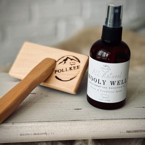 May include: A brown glass bottle of "Wooly Well" restorative sheepskin spray, 4 oz / 120ml, with a black cap. A wooden brush and a wooden block with the "Follkee" logo are also present. The items are on a light gray surface.