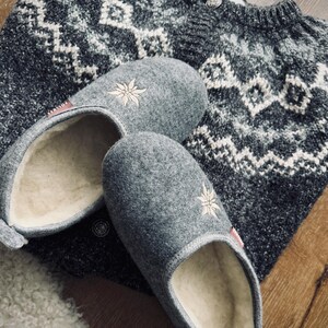 Pode incluir: Um par de pantufas de l&atilde; cinzas com flores brancas bordadas. As pantufas t&ecirc;m um forro de l&atilde; branca e est&atilde;o em cima de um su&eacute;ter com padr&atilde;o cinza e branco.