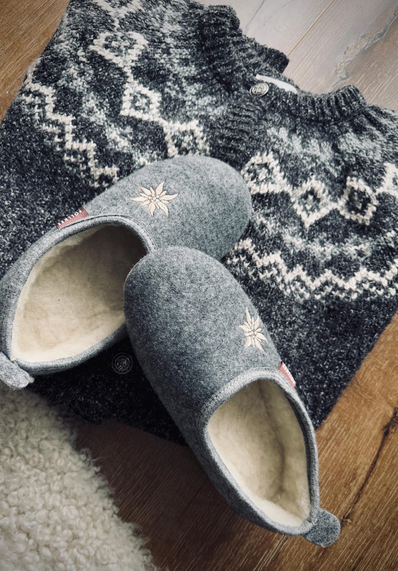 Pode incluir: Um par de pantufas de l&atilde; cinzas com flores brancas bordadas. As pantufas t&ecirc;m um forro de l&atilde; branca e est&atilde;o em cima de um su&eacute;ter com padr&atilde;o cinza e branco.