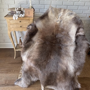 May include: A large, natural reindeer hide rug featuring a thick, dense coat with variegated shades of brown, gray, and cream. The fur has a soft, textured appearance with a multi-tonal color palette, showcasing the organic shape and natural edges.