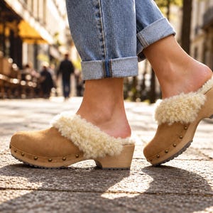 May include: Tan suede clogs featuring a thick, cream-colored shearling lining around the collar. The footwear has a light-colored wooden sole with a row of small, evenly spaced metal studs along the side and a textured dark gray rubber outsole.