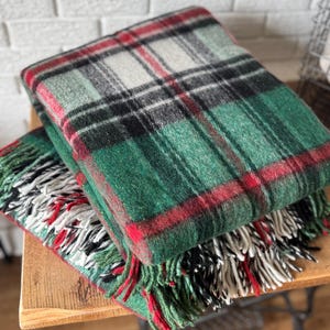May include: Two folded plaid blankets in shades of green, red, black, and white. The blankets have a fringed edge. The blankets are stacked on top of each other on a wooden surface.
