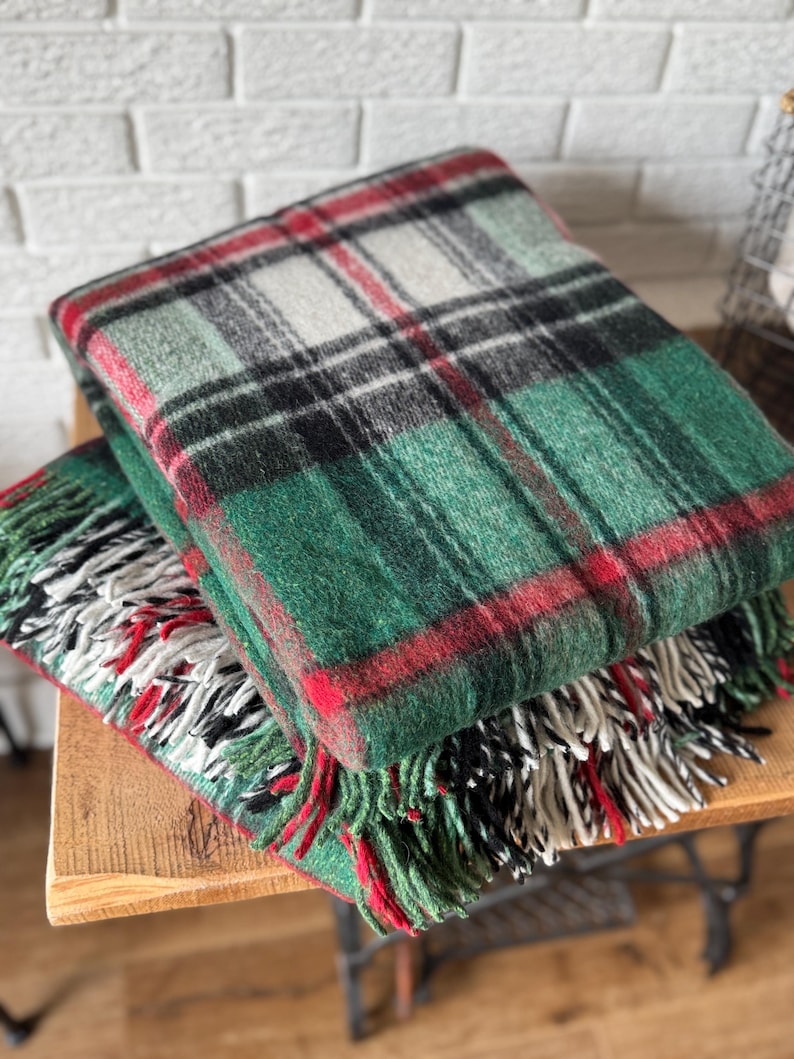 May include: Two folded plaid blankets in shades of green, red, black, and white. The blankets have a fringed edge. The blankets are stacked on top of each other on a wooden surface.