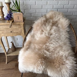 May include: A light beige sheepskin rug with long, soft fibres, draped over a woven chair. The rug has a natural, organic texture. The chair is made of brown wicker and has a rounded back. The scene is set against a white brick wall.