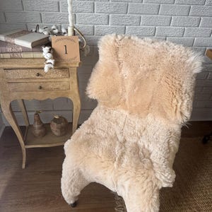 May include: A plush, cream-colored sheepskin rug draped over a chair. The thick, textured fur features a natural, shaggy appearance with soft, warm tones. The rug covers the seat and backrest of the chair, showcasing a dense, fluffy pile.