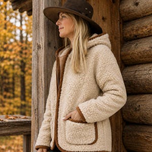May include: A cream-colored sherpa fleece hooded jacket with brown trim, long sleeves, and two front patch pockets. The jacket features a textured, fuzzy exterior and is worn with a dark brown wide-brimmed hat and dark-colored pants.