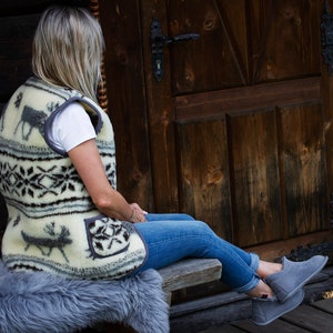 May include: A woman wearing a cream-coloured vest with a black and white geometric pattern and a reindeer design. The vest has a pocket on the left side. She is sitting on a bench with a fluffy grey rug.