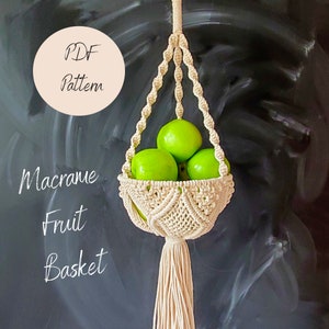 May include: A white macrame fruit basket with a hanging loop and a long tassel. The basket is filled with green apples. The text "PDF Pattern" is written in a circle on the left side of the image. The text "Macrame Fruit Basket" is written below the basket.
