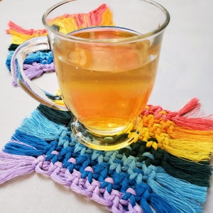 May include: A glass of tea on a colorful crocheted coaster with rainbow fringe. The coaster is made of different colored yarn and has a woven texture.