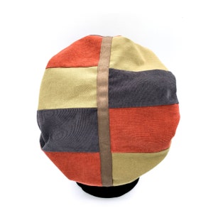 Könnte beinhalten: Ein farbenfroher Cordhut mit Patchwork-Design. Er hat horizontale Streifen in Orange, Grau und Beige mit einem vertikalen braunen Riemen. Der Hut ist vor einem weißen Hintergrund platziert.