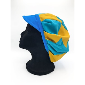 Könnte beinhalten: Ein farbenfroher Kopfschmuck mit Visier. Der Hut hat ein Patchwork-Design aus türkisblauem, gelbem und blauem Stoff. Das Visier und die Zierleiste sind einfarbig blau. Der Hut wird auf einem schwarzen Schaufensterkopf präsentiert.