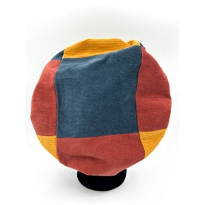 Könnte beinhalten: Ein farbenfroher Cordhut im Patchwork-Design. Der Hut hat Einsätze in Rot, Marineblau und Gelbgold. Der Hut wird auf einem schwarzen Ständer vor weißem Hintergrund präsentiert.