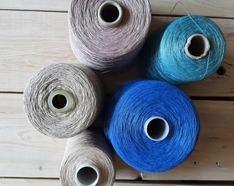 May include: Five spools of yarn in different colours and textures. The spools are arranged in a circle on a wooden surface. The colours are beige, light blue, dark blue, and a darker shade of beige.
