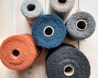 Puede incluir: Seis carretes de hilo de diferentes colores y texturas. Los carretes están dispuestos en círculo sobre una superficie de madera clara. Los colores son gris, azul, naranja, negro y beige.