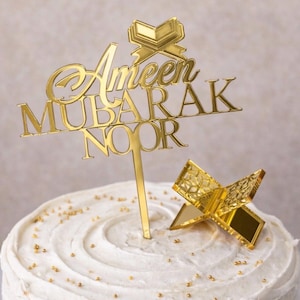 May include: A gold cake topper with the words "Ameen Mubarak Noor" and a small open book design. The topper is placed on a white frosted cake with gold sprinkles.