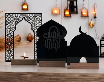 Op de afbeelding: Drie zwarte houten decoratieve stukken met ingewikkelde ontwerpen. Het stuk aan de linkerkant is een raamvormig frame met een geometrisch patroon. Het middelste stuk is een koepel met de Arabische tekst "Eid Mubarak" erop. Het stuk aan de rechterkant is een koepel met een halve maan erop. Alle drie stukken staan op een houten oppervlak.