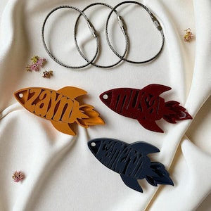 May include: Three metal keychains and three rocket-shaped keychains in orange, burgundy, and navy blue. The orange keychain has the name "Zayn" cut out, the burgundy keychain has the name "Musa", and the navy blue keychain has the name "Waliyah".