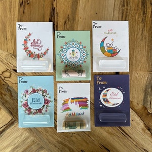 May include: Five colorful gift card holders with a clear plastic window to insert a gift card or cash. Each card holder features a different design with a crescent moon and Islamic greetings in Arabic script. The greetings include "Eid Mubarak" and "Eid al-Fitr".