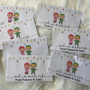 May include: A set of six white cards with a cartoon illustration of two children holding hands. The cards have a string of colorful lights at the top and the text "eid mubarak" with a heart symbol. Some cards have the text "From Yameen & Sobia".