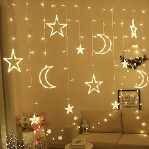 May include: Warm white string lights with star and crescent moon shapes. The lights are hanging in a curtain-like arrangement, creating a soft, ambient glow. A decorative wreath and framed artwork are also visible.