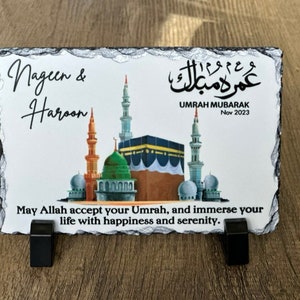 May include: A black slate plaque with a white background featuring a colorful illustration of the Kaaba and other mosques. The text "Nageen & Haroon" and "Umrah Mubarak Nov 2023" is printed on the plaque. The plaque is on a wooden surface.