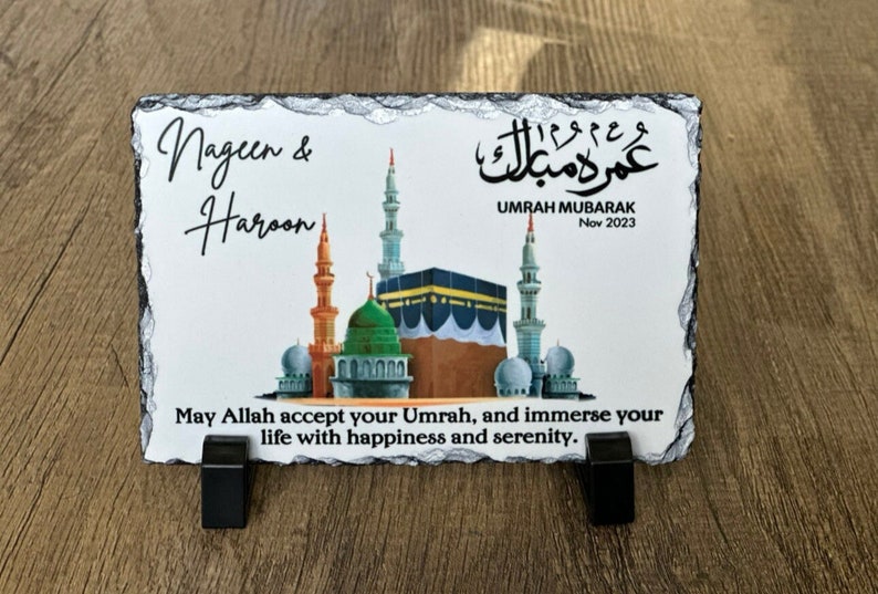 May include: A black slate plaque with a white background featuring a colorful illustration of the Kaaba and other mosques. The text "Nageen & Haroon" and "Umrah Mubarak Nov 2023" is printed on the plaque. The plaque is on a wooden surface.