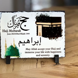May include: White slate plaque with a black and gold image of the Kaaba, a green mosque, and Arabic calligraphy. The text reads "Hajj Mubarak June 2023/Dhul Hijjah 1444" and "May Allah accept your Hajj and immerse your life with happiness and serenity."
